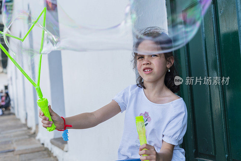 拉丁女孩在Villa de Leyva的广场上玩起泡器制造巨大的气泡，同时她在微风中跑步和玩耍